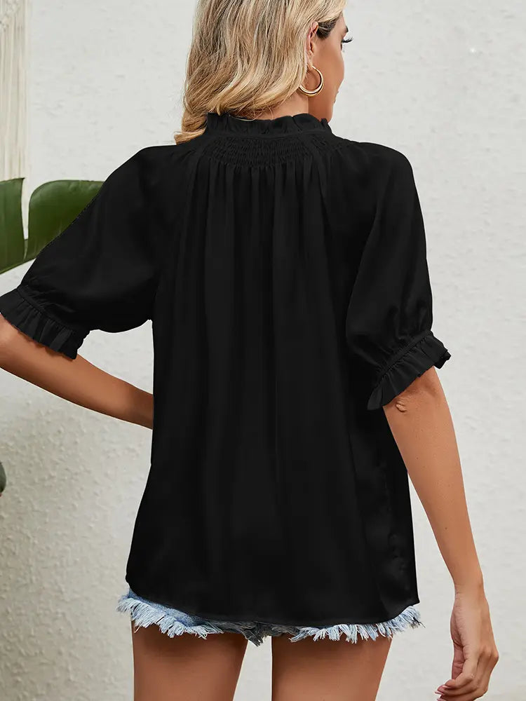 Woman wearing a black blouse with rolled-up sleeves and denim shorts against a white wall.